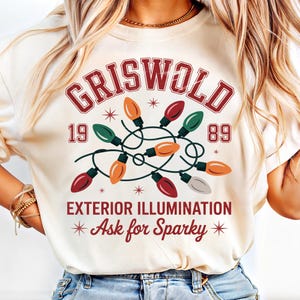 May include: Cream-colored sweatshirt featuring the word "GRISWOLD" in a red, vintage-style font, with the numbers "19" and "89". A string of colorful Christmas lights is in the center, with the text "EXTERIOR ILLUMINATION" and "Ask for Sparky".