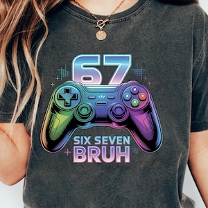 Könnte beinhalten: Dunkelgraues T-Shirt mit einem farbenfrohen Videospiel-Controller-Motiv und der Zahl 67 in einem Farbverlauf. Der Text "SIX SEVEN BRUH" befindet sich unter dem Controller.