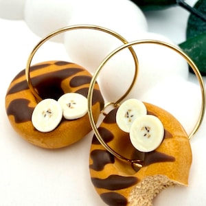 Könnte beinhalten: Ein Paar goldene Creolen-Ohrringe mit einem Miniatur-Donut-Design. Die Donuts haben ein braunes und beiges Muster, Schokoladenglasur und weiße Bananenscheiben. Ein Donut hat einen Biss.