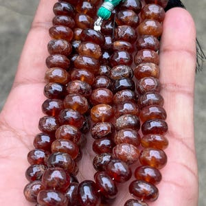 May include: Strands of polished, reddish-brown gemstone beads. The beads are oval-shaped and strung together, with a green and black tassel at the top. The beads are held in a hand, showcasing their color and texture.