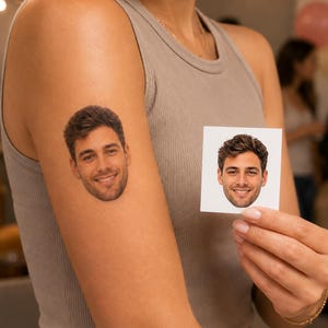Può includere: Un tatuaggio temporaneo che raffigura il ritratto realistico di un uomo sorridente con capelli castano scuro e una barba leggera, applicato sulla parte superiore del braccio. Viene tenuta una stampa quadrata su carta dello stesso ritratto.