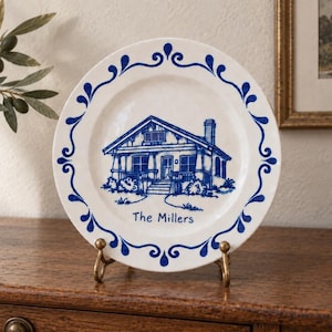 May include: A white ceramic plate featuring a blue illustration of a house in the centre and the text "The Millers" below it. The rim of the plate is decorated with a blue scrollwork pattern. The plate is displayed on a gold metal stand.