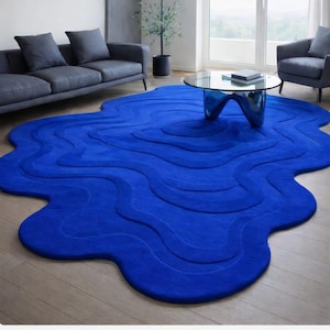 May include: A vibrant blue, abstract-shaped rug with a wavy design dominates a living room. The rug's unique form and color create a focal point, complementing the gray sofa, chairs, and glass coffee table. The rug is made of a soft material.