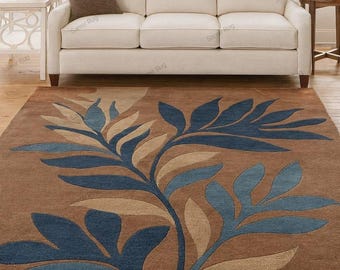 May include: A brown area rug with a large, stylized leaf design in shades of blue and tan. The rug is placed in front of a cream-colored sofa, creating a warm and inviting living space. The rug's dimensions are not visible.