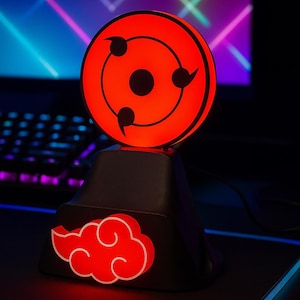 Peut inclure: Lampe de bureau rouge et noire sur le thème de l'anime. La lampe présente un motif d'œil rougeoyant avec trois tomoe noirs. La base présente un motif de nuage rouge. La lampe est sur une surface noire.