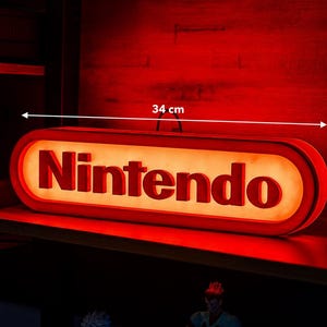 Può includere: Un'insegna rettangolare illuminata Nintendo con bordi arrotondati, caratterizzata dal logo rosso Nintendo in rilievo su uno sfondo arancione brillante. L'insegna misura 34 cm di larghezza e 10-12 cm di altezza, come indicato dalle frecce.