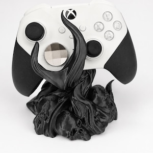 Peut inclure: Une manette Xbox blanche et noire repose sur un support noir stylisé. Le support présente un design détaillé avec des crânes et des formes organiques. La manette a des poignées et des boutons noirs, avec le logo Xbox visible.