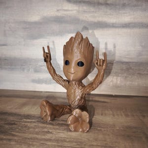 May include: A brown figurine of a character with a tree-like appearance, seated with arms raised in a rock-and-roll gesture. The figure has a smiling face and is set against a wooden background.