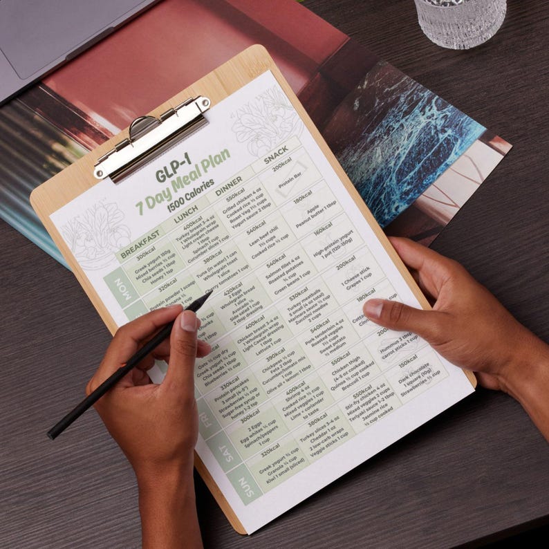 May include: A wooden clipboard with a printed 7-day meal plan, held by a person. The plan includes daily calorie targets and meal suggestions. A pen is held over the plan, ready to write. The clipboard has a metal clip at the top.