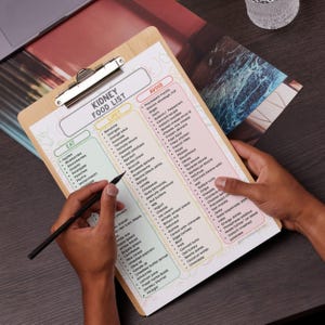 May include: A wooden clipboard with a silver clip holds a printed "Kidney Food List." The list categorizes foods into "Eat," "Limit," and "Avoid" sections. A hand holds a black pen, poised to write on the list. The clipboard is on a dark surface.