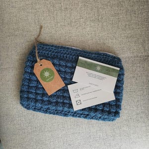 Peut inclure: Une pochette bleue crochetée avec une fermeture éclair. Une étiquette marron avec un logo vert et le texte "HAZE STUDIOS" est attachée. Deux cartes blanches avec des instructions d'entretien et du texte sont également présentes.