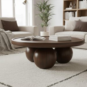 Op de afbeelding: Een ronde, donkerbruine salontafel met drie bolvormige poten. Op de tafel liggen boeken en een open boek. De tafel staat in een woonkamer met een wit tapijt en een bank.