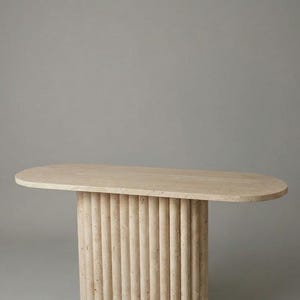 Può includere: Tavolo ovale beige con base cilindrica. La base è composta da colonne verticali e arrotondate. Il tavolo è realizzato in pietra di colore chiaro, su sfondo grigio.