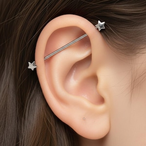 May include: A silver industrial barbell earring with star-shaped ends. The earring is inserted through the ear cartilage. The image shows a close-up of the ear, highlighting the jewelry's design and placement. The earring is a popular body modification.