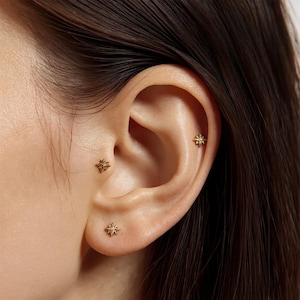 May include: Close-up of an ear with three small, gold-colored star-shaped earrings. The earrings are placed in different locations on the ear. The skin tone is light, and the hair is dark brown.