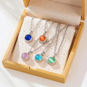 Peut inclure: Six colliers argentés avec des pendentifs ronds et colorés. Les pierres sont bleues, orange, roses, turquoise et vertes. Les colliers sont présentés dans un coffret en bois avec un intérieur crème.