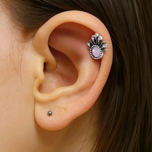 May include: A silver-colored ear piercing featuring a crown design with a light pink oval-shaped stone. The piercing is set against the backdrop of a person's ear, with a small, silver stud visible in the earlobe.