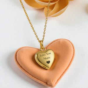 May include: A gold-colored heart-shaped pendant necklace with the words "Love You Mum" and a small black heart engraved on the front. The pendant hangs from a delicate chain and rests on a peach-colored heart-shaped cushion.
