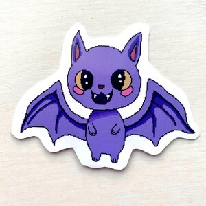 Può includere: Un adesivo a forma di pipistrello viola con ali e corpo pixelati. Il pipistrello ha grandi occhi rotondi, guance rosa e una bocca sorridente con piccole zanne. L'adesivo è su sfondo bianco.