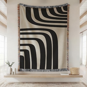 May include: A large, rectangular throw blanket with a cream background and bold black curved lines. The blanket has a fringed edge with a mix of colors. The design is modern and geometric, suitable for a living room or bedroom.