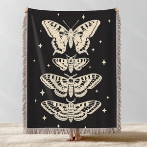 May include: Black throw blanket with cream-coloured butterfly and moth illustrations. The insects are arranged vertically with star accents. The blanket has a fringed edge.