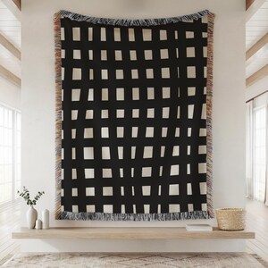 May include: Black and white woven throw blanket with a grid pattern of irregular off-white rectangles. The blanket has a fringed edge with a mix of colors. The throw is displayed on a wall, adding a modern touch to the room.