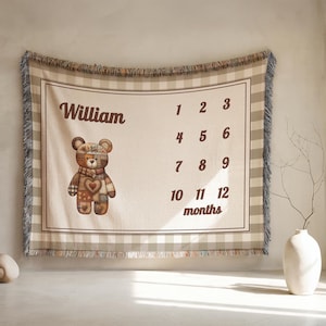 May include: A beige and brown woven baby milestone blanket with a teddy bear illustration and the name "William". The blanket features a gingham border and fringe trim, with numbers 1-12 for monthly milestones.