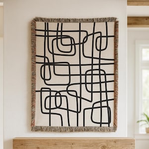 Op de afbeelding: Een geweven sprei met een crèmekleurige achtergrond en een zwart abstract geometrisch ontwerp. De sprei heeft een gefranjerde rand in bruin- en roodtinten. Perfect om een moderne touch aan elke woonruimte toe te voegen.