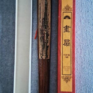 May include: A closed, brown folding fan with gold accents, a white box, and a red box with gold text. The red box has the words "PAINTING FANS" in English. The fan is made of thin wooden slats.