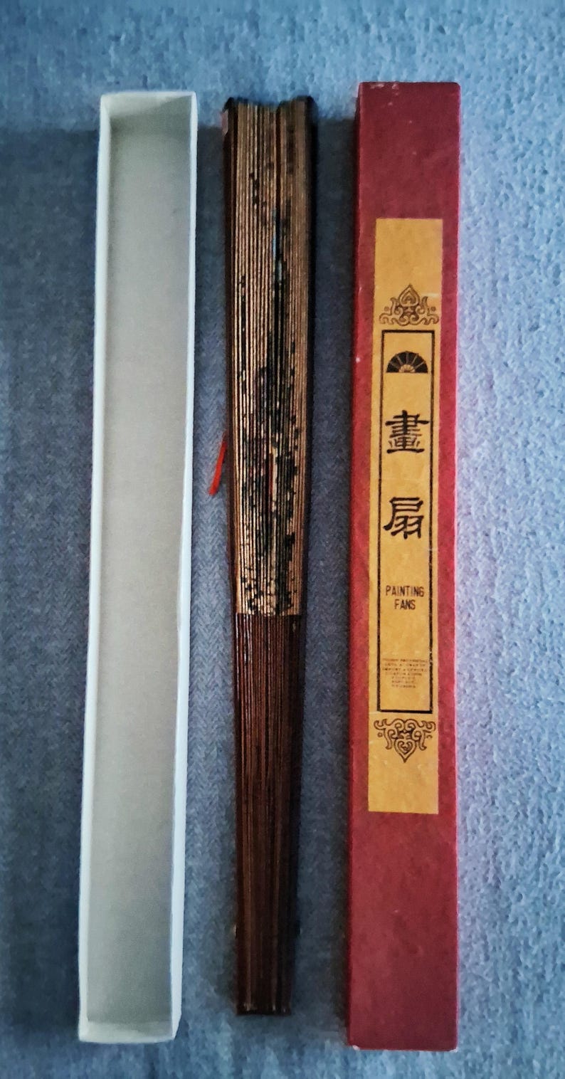 May include: A closed, brown folding fan with gold accents, a white box, and a red box with gold text. The red box has the words "PAINTING FANS" in English. The fan is made of thin wooden slats.