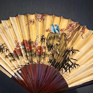 May include: Hand-painted fan with a yellow background, featuring red flowers, bamboo, and two blue birds perched on a rock. The fan's wooden handle is a deep burgundy color, and the fan is open.
