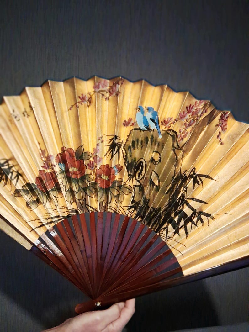 May include: Hand-painted fan with a yellow background, featuring red flowers, bamboo, and two blue birds perched on a rock. The fan's wooden handle is a deep burgundy color, and the fan is open.