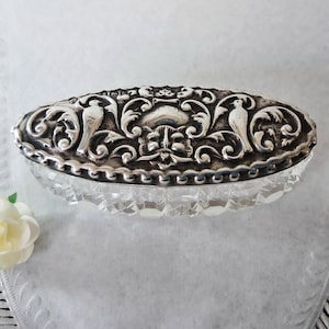 May include: An oval silver trinket box with a clear, faceted base. The lid is decorated with detailed scrollwork, birds, and floral patterns. The box is presented with artificial flowers.