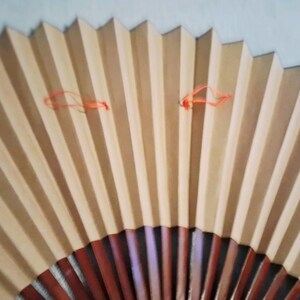 May include: A partially open, pleated paper fan with alternating light brown and beige vertical stripes. The fan's base is a darker reddish-brown, and it has two orange strings attached near the top.