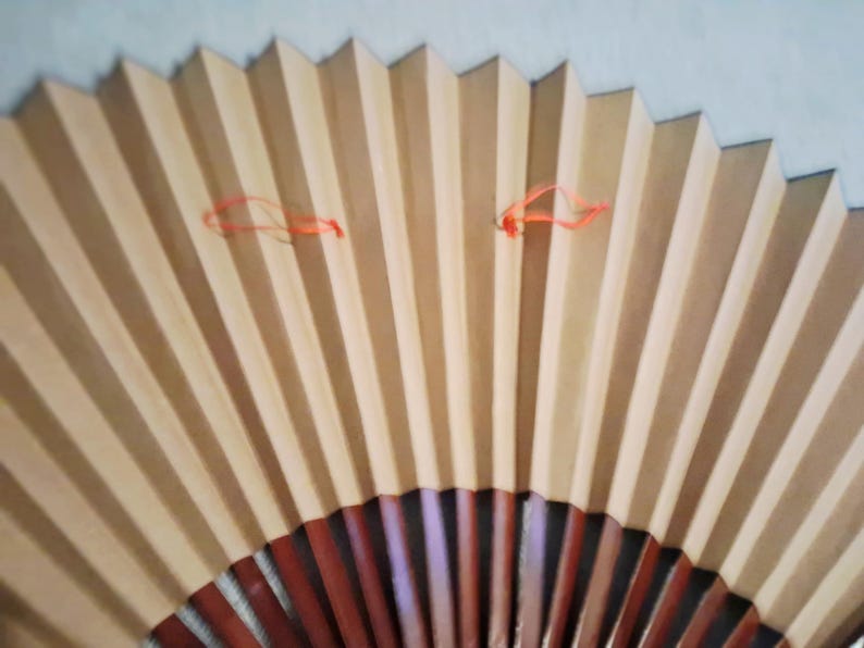 May include: A partially open, pleated paper fan with alternating light brown and beige vertical stripes. The fan's base is a darker reddish-brown, and it has two orange strings attached near the top.