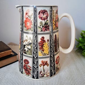 May include: A ceramic jug featuring a grid of rectangular panels, each displaying a different botanical illustration of flowers including petunias, violas, and wallflowers. The panels are separated by black borders with white leaf motifs.