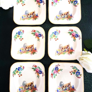 May include: A set of six square ceramic saucers with rounded corners, featuring a central illustration of a thatched-roof cottage surrounded by colourful flowers. Each saucer has a gold-trimmed edge and floral accents in the top corners.
