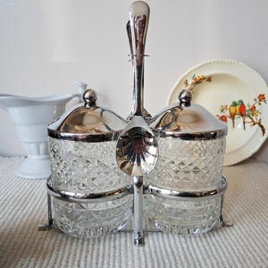 May include: A vintage condiment set with two glass jars, chrome lids, and spoons. The jars have a diamond pattern and sit in a chrome holder. A white gravy boat and a decorative plate with birds are in the background.