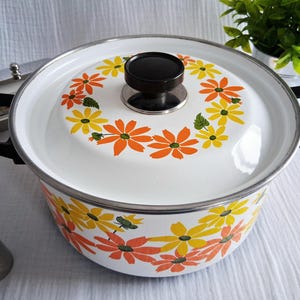 May include: White enamel pot with a lid featuring a floral design in orange and yellow. The pot has black handles and a black knob on the lid. A silver rim and accents complete the look. A vintage kitchenware item.