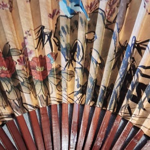 May include: A hand fan with a painted design featuring birds, flowers, and foliage in shades of red, blue, and brown. The fan's ribs are made of dark brown wood. The fan is open, displaying the artwork.