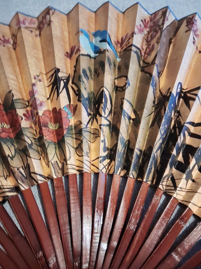 May include: A hand fan with a painted design featuring birds, flowers, and foliage in shades of red, blue, and brown. The fan's ribs are made of dark brown wood. The fan is open, displaying the artwork.