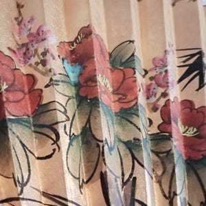 May include: A close-up of a hand fan with a floral design. The fan features red flowers with green leaves and pink blossoms against a beige background. The fan is made of paper and has a pleated design.