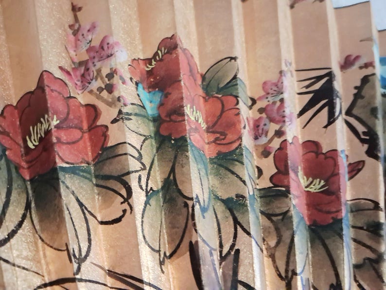 May include: A close-up of a hand fan with a floral design. The fan features red flowers with green leaves and pink blossoms against a beige background. The fan is made of paper and has a pleated design.