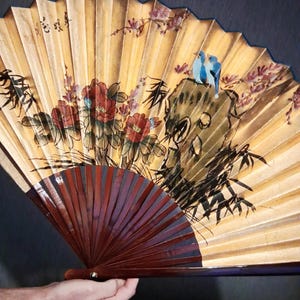 May include: A hand-held folding fan with a gold background and a dark brown handle. The fan features a painted design of red flowers, bamboo, and two blue birds perched on a rock. The fan is held open, displaying its full design.