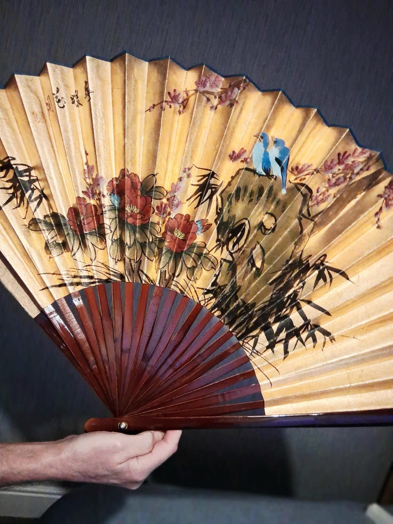 May include: A hand-held folding fan with a gold background and a dark brown handle. The fan features a painted design of red flowers, bamboo, and two blue birds perched on a rock. The fan is held open, displaying its full design.