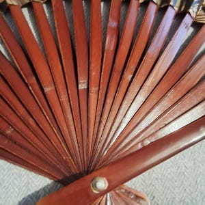 May include: A wooden hand fan with a dark reddish-brown frame and a decorative top section. The fan's frame is made of thin, evenly spaced wooden slats. The top section features a patterned design with shades of beige, brown, and black.