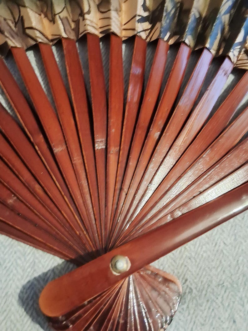 May include: A wooden hand fan with a dark reddish-brown frame and a decorative top section. The fan's frame is made of thin, evenly spaced wooden slats. The top section features a patterned design with shades of beige, brown, and black.