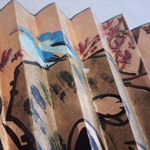 May include: Close-up of a decorative folding fan with a painted design. The fan features a tan background with a blue bird and pink floral accents. Black brushstrokes outline the design, creating a traditional Asian aesthetic.