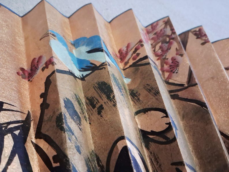 May include: Close-up of a decorative folding fan with a painted design. The fan features a tan background with a blue bird and pink floral accents. Black brushstrokes outline the design, creating a traditional Asian aesthetic.