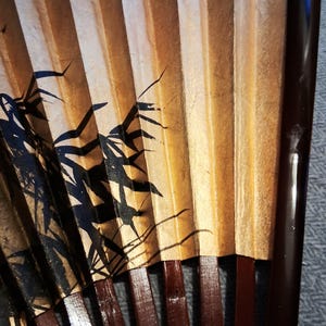 May include: A close-up of a traditional hand fan. The fan features a bamboo design in black ink on a gold and tan background. The ribs are a dark brown color.